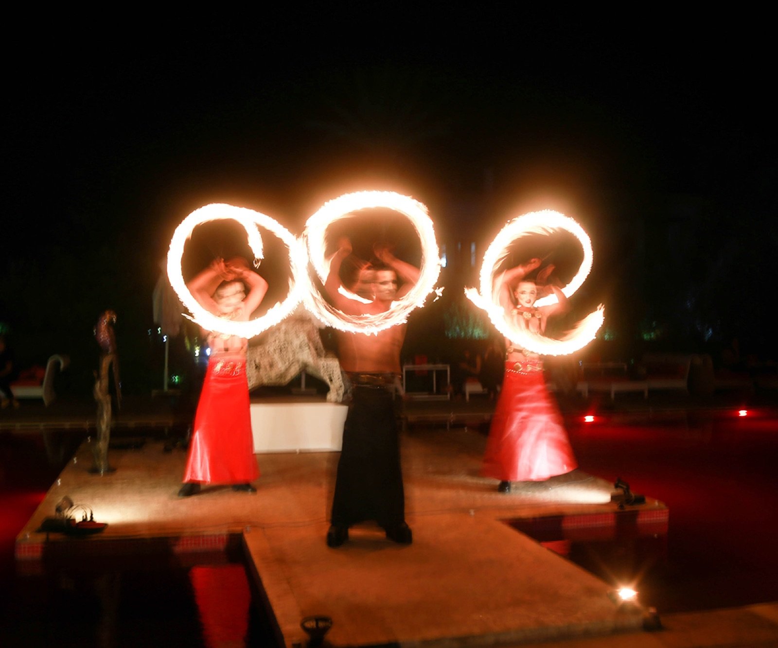 Three fire performers spinning fire poi on a poolside stage
